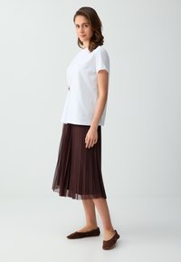 Jeune femme debout de profil portant un haut blanc à manches courtes, une jupe midi plissée marron et des chaussures plates marron sur un fond uni.