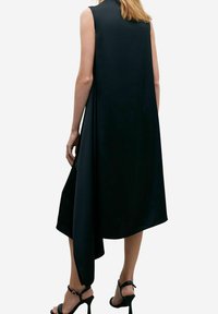 Robe noire sans manches avec un ourlet asymétrique, texture lisse et design fluide. Portée avec des talons hauts noirs à lanières.