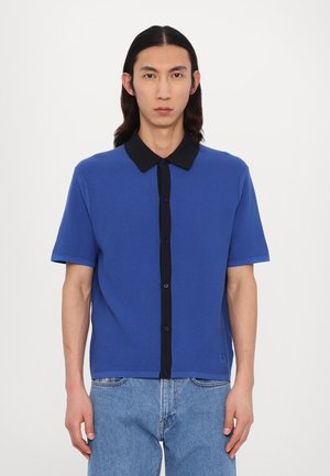 Short-sleeve polo shirt in blue, featuring a textured fabric, black collar, and buttoned front. Worn with light blue denim jeans.