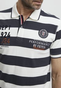 Polo a rayas en navy y blanco que presenta un logotipo bordado, una tapeta con botones y un texto decorativo en naranja en el pecho.