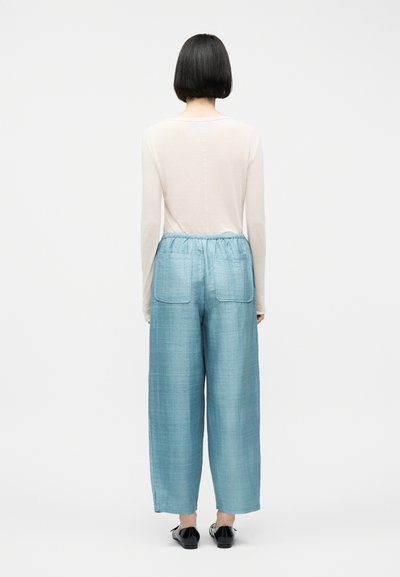 Γυναίκα με κοντά μαύρα μαλλιά στέκεται γυρισμένη με την πλάτη φορώντας ένα διάφανο λευκό μπλουζάκι με μακριά μανίκια, ανοιχτόχρωμο μπλε παντελόνι με φαρδιά μπατζάκια και τσέπες στο πίσω μέρος, και μαύρα παπούτσια.