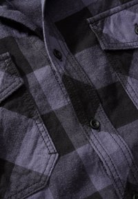 Paarse en zwarte geruite flanellen shirt met knoopsluitingen en twee borstzakken met puntflappen; de stof heeft een zachte textuur.