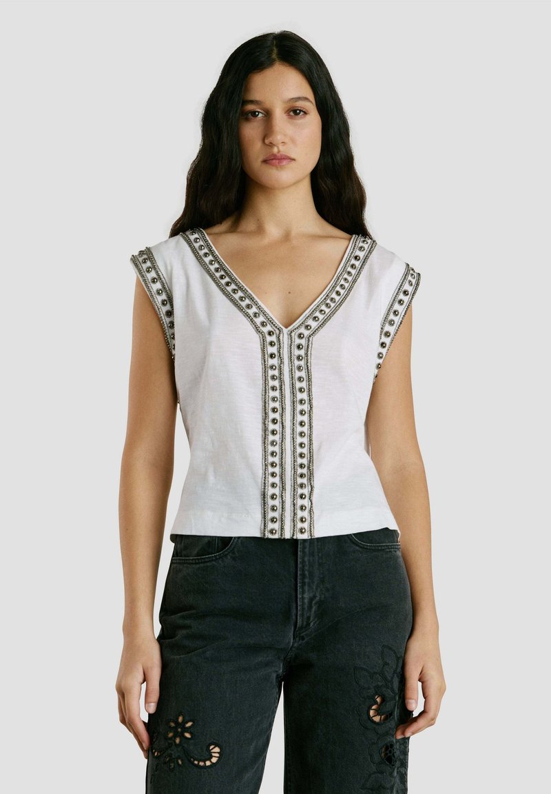 Kvinde med langt mørkt hår iført en ærmeløs hvid bluse med perledetaljer og sorte jeans med blomstermønstrede udskæringer, stående foran en grå baggrund.