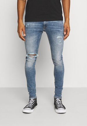 Ben iført falmede blå, slidte skinny jeans og sorte høje sneakers med hvide snørebånd og såler, stående på gråt gulv.