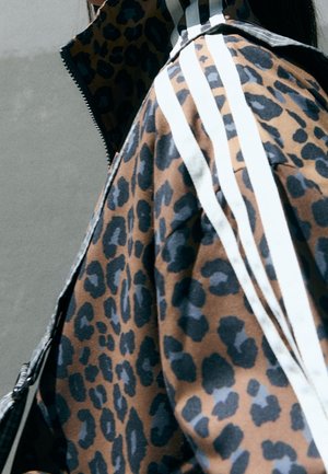Person iført en brun og sort leopardprintjakke med hvide tre-stribede detaljer på skulderen mod en ensfarvet grå baggrund.