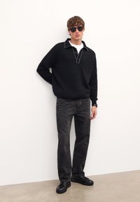 Zwarte trui met een kraag en witte stiksels, gecombineerd met donkere jeans en zwarte sneakers. Accessoires zijn donkere zonnebrillen.