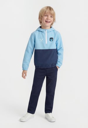 Garçon souriant debout avec une main dans la poche, portant une veste à capuche bleu et marine, un pantalon marine et des baskets blanches sur fond blanc.