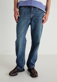 Jeans de denim azul com um corte descontraído e um ligeiro desbotado, design de cinco bolsos e costura em contraste, usados com ténis escuros.