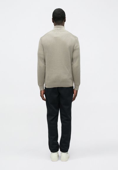 Pull en tricot beige clair, avec une texture côtelée et une coupe ajustée, associé à un pantalon de jogging foncé et des baskets blanches.