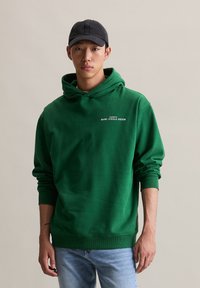 Jonge man met een zwarte pet, groene hoodie met de tekst "MARC O'POLO DENIM" en lichtblauwe jeans, staande tegen een effen beige achtergrond.