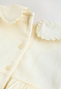 Vêtement de couleur jaune clair avec un col festonné, une fermeture à boutons et de délicates plis en bas. Fabriqué en tissu doux avec une texture lisse.