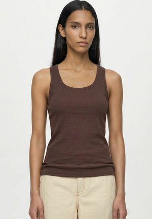Jeune femme aux longs cheveux foncés, portant un top marron sans manches et un pantalon beige, debout devant un fond clair uni.