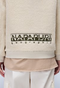 Κάπα φλις σε μπεζ απόχρωση με υφή, διαθέτει λογότυπο σε σκούρο πράσινο χρώμα στην πλάτη, περιτοιχισμένο από μια κρέμα απόχρωση στο τελείωμα.