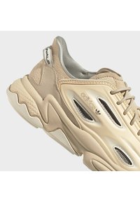 Béžová teniska Adidas s tlustou podrážkou, síťovanými ventilačními panely, semišovou patou a označeným poutkem, zobrazená z bočního úhlu.