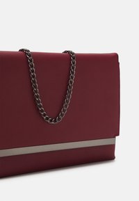 Pochette bordeaux avec une texture mate, dotée d'une bandoulière en chaîne argentée et d'un accent métallique horizontal sur le devant.