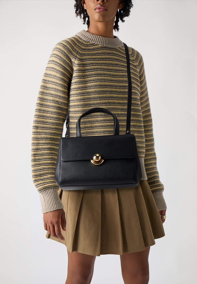Sac à main en cuir noir avec une forme structurée, des accents en métal doré et une poignée supérieure, suspendu à une sangle noire.