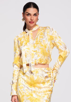 Femme aux cheveux foncés portant un haut à manches longues jaune et blanc à motifs floraux et une jupe assortie, posant avec une main sur la hanche devant un fond uni.