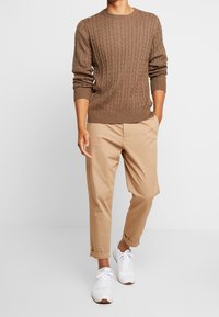 Pull en tricot beige à col rond, associé à un chino beige retroussé et des baskets blanches avec des accents marron.