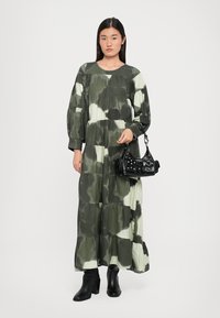 Pitkä, kerroksittain leikattu mekko, jossa on camouflage-kuvio vihreän ja kerman sävyissä; siinä on pitkät hihat, pyöreä pääntie ja valuva kangas.