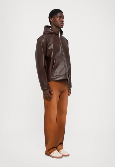 Mand står iført en mørkebrun læderjacket med hætte, brændt orange bukser og beige sneakers foran en ensfarvet lys baggrund.