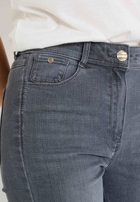 Jean de mezclilla gris con una cintura alta, bolsillos frontales y detalles en tono dorado en la cinturilla y en el bolsillo. Textura suave.