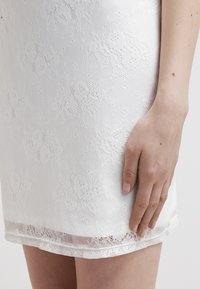 Bílé krajkové šaty s květinovými vzory, texturovaným tkaním a zoubkovaným lemem. Ruka jemně spočívá na látce, což zdůrazňuje její detaily.