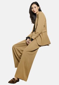 Tan op maat gemaakte blazer met een gelaagd ontwerp, gecombineerd met brede tan broeken. Getextureerde loafers met luipaardprint. Zwarte blouse eronder.