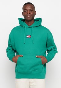 Hanorac verde cu glugă, realizat dintr-un material texturat, având o buzunar frontal tip kangaroo și un mic patch cu logo în culori contrastante.