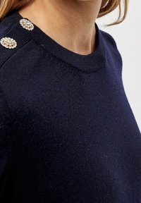 Marinblå stickad tröja med rund halsringning, slät textur och tre dekorativa juvelknappar på axeln.