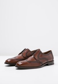Scarpe in pelle marrone con dettagli brogue, caratterizzate da una punta affusolata, design con lacci e una superficie liscia. Suole piatte e sottili accenti rossi.