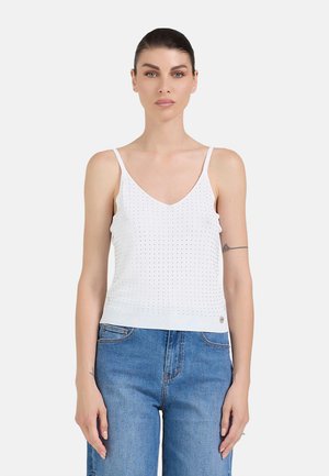 Donna con capelli scuri, con un top bianco senza maniche e jeans blu, in piedi di fronte a uno sfondo chiaro e uniforme.
