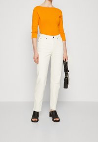 Oranges Langarmoberteil, weiße hoch taillierte Jeans, schwarze Blockabsatz-Mules und eine schwarze Handtasche. Einfaches Design und kräftige Farben.
