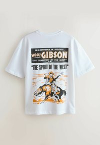 Fehér, rövid ujjú póló, amely egy vintage poszter dizájnt ábrázol: egy cowboy lovon, a "Hoot Gibson" és a "The Spirit of the West" feliratokkal.