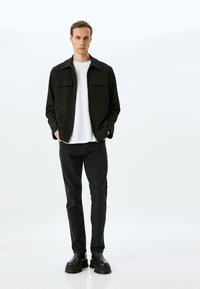 Chaqueta negra con cremallera y dos bolsillos en el pecho, sobre una camiseta blanca. Combinada con jeans oscuros y botas negras gruesas. Tejido suave, corte entallado.