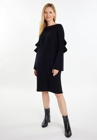 Abito nero a maglia con maniche a volant, design al ginocchio e taglio dritto. Abbinato a stivali neri alla caviglia. Texture liscia, stile minimalista.