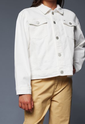 Persona con chaqueta vaquera blanca abotonada y bolsillos con solapa, y pantalones beige, de pie contra un fondo gris.