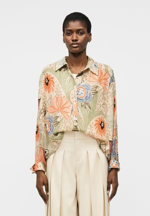 Kobieta ubrana w luźną, kwiecistą koszulę zapinaną na guziki z pomarańczowymi, niebieskimi i zielonymi wzorami, wpuszczoną w kremowe, plisowane spodnie z wysokim stanem.