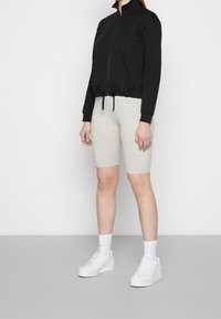 Zwarte zip-up jack met hoge kraag, elastische trekkoord onderaan, gecombineerd met lichtgrijze knielange shorts. Witte sneakers en enkelkousen maken de look compleet.
