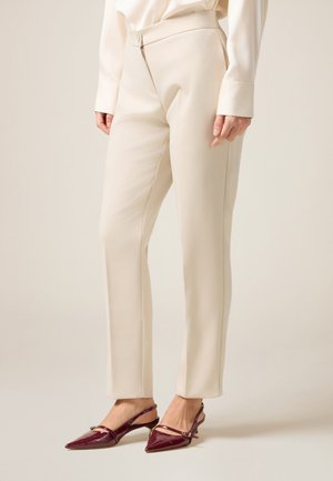 Beige maßgeschneiderte Hosen mit glatter Textur, featuring einen Reißverschluss und Knopfverschluss vorne. Getragen mit burgunderfarbenen, glänzenden Slingback-Absätzen.