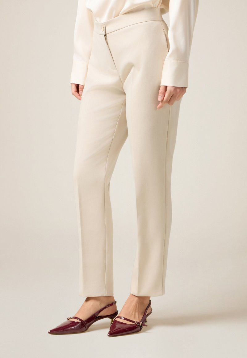 Beige maßgeschneiderte Hosen mit glatter Textur, featuring einen Reißverschluss und Knopfverschluss vorne. Getragen mit burgunderfarbenen, glänzenden Slingback-Absätzen.