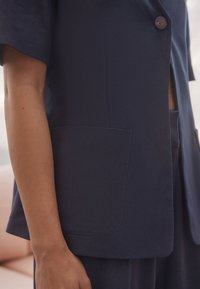 Blazer marine en tissu texturé avec manches courtes, fermeture à un bouton et une poche plaquée sur le côté gauche.