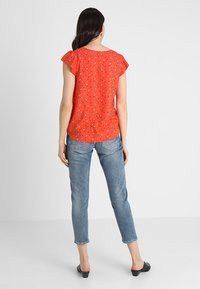 Rode blouse met korte mouwen met kleine blauwe en witte bloemmotieven, gecombineerd met lichtblauwe enkelbroeken en zwarte block-heeled schoenen.