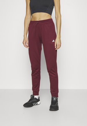 Persoon die kastanjebruine Adidas joggers, zwart crop top, witte sokken en zwarte Adidas sneakers draagt, staand tegen een egale witte achtergrond.