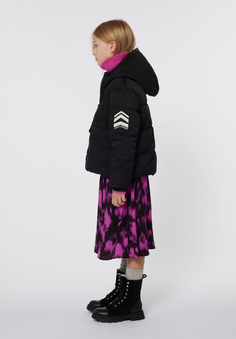 Zwarte pufferjack met capuchon, gestreepte patch op de mouw, gedragen met een levendig roze tie-dye jurk en zwarte veterschoenen.