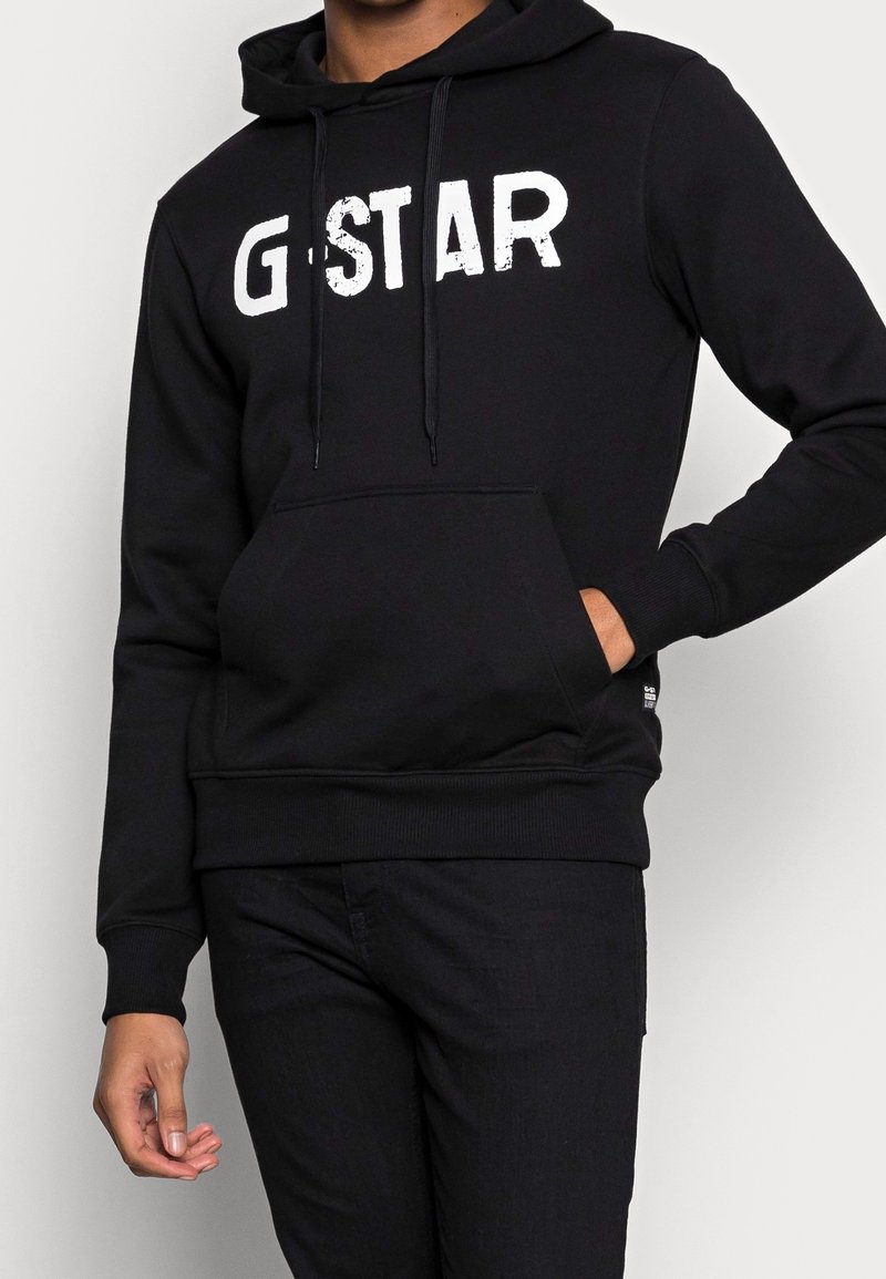 Sudadera negra hecha de una tela suave, con un bolsillo frontal y el texto "G-STAR" en blanco y desgastado. Puños y dobladillo acanalados para un mejor ajuste.