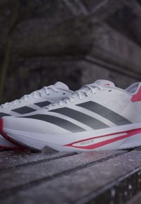 Hvide atletiksneakers med sorte striber og lyserøde accenter, hvilende på en træoverflade udendørs ved skumring.