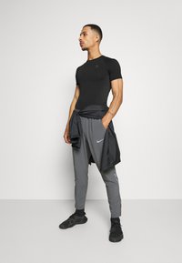Schwarzes kurzärmliges Sportshirt mit strukturiertem Design, kombiniert mit grauen schmal zulaufenden Hosen mit Nike-Logo und schwarzen Sportschuhen.