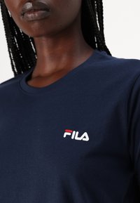Námornícky modré bavlnené tričko s krátkymi rukávmi, s bielym a červeným vyšívaným logom FILA na ľavej strane hrudníka. Hladký povrch.