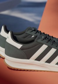 Baskets vert foncé et blanc avec trois bandes blanches et semelle « Cloudfoam » sur une surface orange avec un fond bleu.