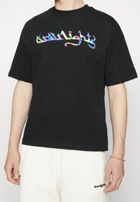 Person in einem schwarzen T-Shirt mit dem Regenbogen-Text "Lowlights" und weißen Hosen mit kleinem schwarzem "Lowlights"-Logo auf dem linken Oberschenkel.